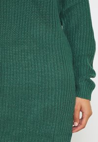 Robe en tricot verte avec un motif texturé et côtelé. Dotée de manches longues et de poignets ajustés, mettant en valeur un tissu doux et confortable.