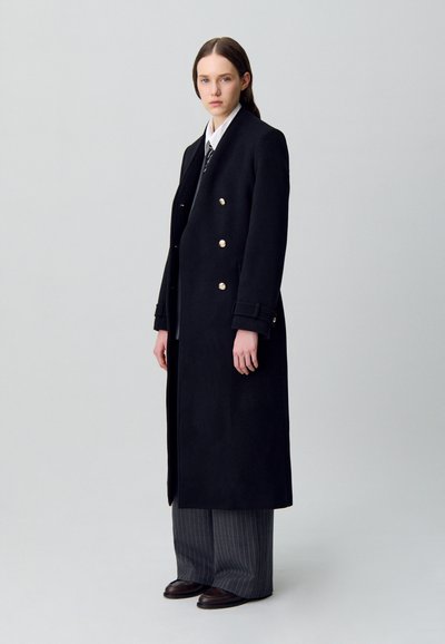 Cappotto lungo nero con bottoni d'argento doppiopetto, vestibilità strutturata e ampi revers, abbinato a pantaloni a righe sottili e scarpe scure.