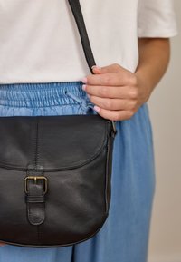 Czarna skórzana torba na ramię z zakrzywioną klapą, ze szczegółem w postaci mosiężnej klamry i regulowanym paskiem, trzymana w dłoni. Fakturowana powierzchnia.