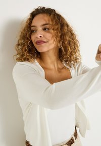 Cardigan blanc au toucher doux, porté sur un haut léger. Le modèle a les cheveux bouclés et une simple ceinture à imprimé léopard à la taille.
