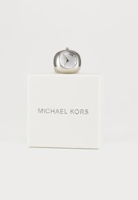 Silberfarbene ovale Uhr mit weißem Zifferblatt, römischen Ziffern und gewölbtem Glas; präsentiert auf einer strukturierten weißen Box mit dem "MICHAEL KORS"-Logo.
