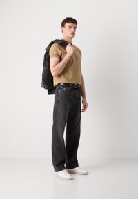 Beige Polo-Shirt mit weißem Besatz, dunkelgraue Jeans aus Denim und weiße Sneakers; Model hält eine schwarze Jeansjacke über der Schulter.
