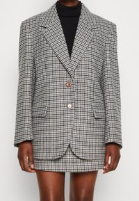 Harmaasta ja mustasta houndstooth-kuviollinen bleiseri, jossa kaksi etutaskua, yksittäinen halkio ja ruskeat napit. Räätälöity istuvuus.