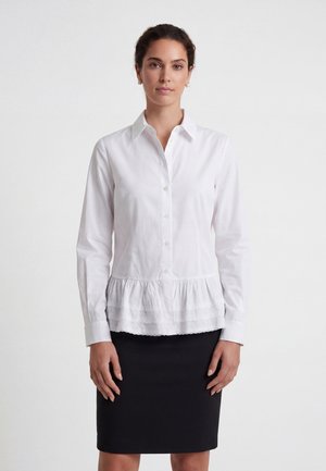 Woman with dark hair in a white buttoned blouse with ruffled hem and a black knee-length skirt standing against a plain white background.