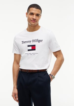 Άνδρας που φοράει λευκό T-shirt Tommy Hilfiger με λογότυπο και κείμενο, σκούρο παντελόνι, καφέ ζώνη και ρολόι χειρός, στέκεται μπροστά σε απλό λευκό φόντο.