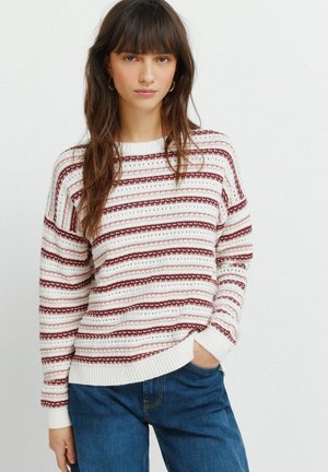 Giovane donna con lunghi capelli castani, indossa un maglione a righe bianche e bordeaux e jeans blu, in piedi con una mano in tasca.