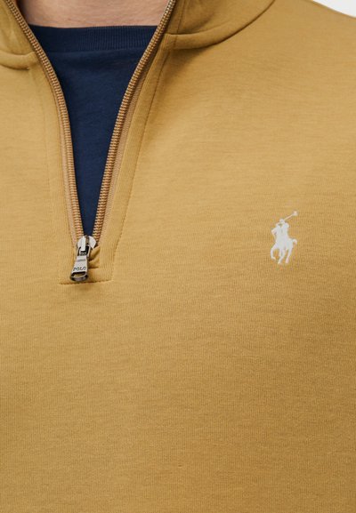 Μπεζ πουλόβερ με φερμουάρ στο ένα τέταρτο μήκους σε ασημί χρώμα και λευκό κεντητό λογότυπο παίκτη πόλο στο στήθος, φορεμένο πάνω από μπλε σκούρο πουκάμισο.