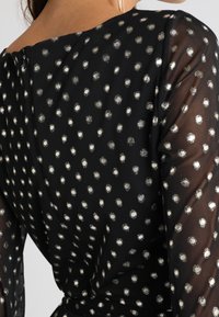 Schwarze Bluse aus Stoff mit metallisch-silbernen Pünktchen, transparenten Ärmeln und einem leichten V-Ausschnitt. Enthält einen Reißverschluss auf der Rückseite.