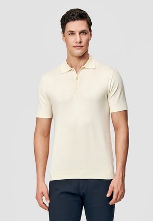 Mężczyzna ubrany w kremową, krótkorękawową koszulkę polo i ciemne spodnie, stoi na tle jednolitego jasnego tła.