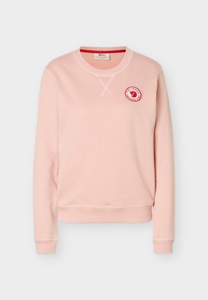 Svetlo roza majica s posadkim ovratnikom, dolgimi rokavi in majhnim okroglim rdeče-belim logotipom Fjällräven Sweden na zgornjem levem delu prsnega koša.