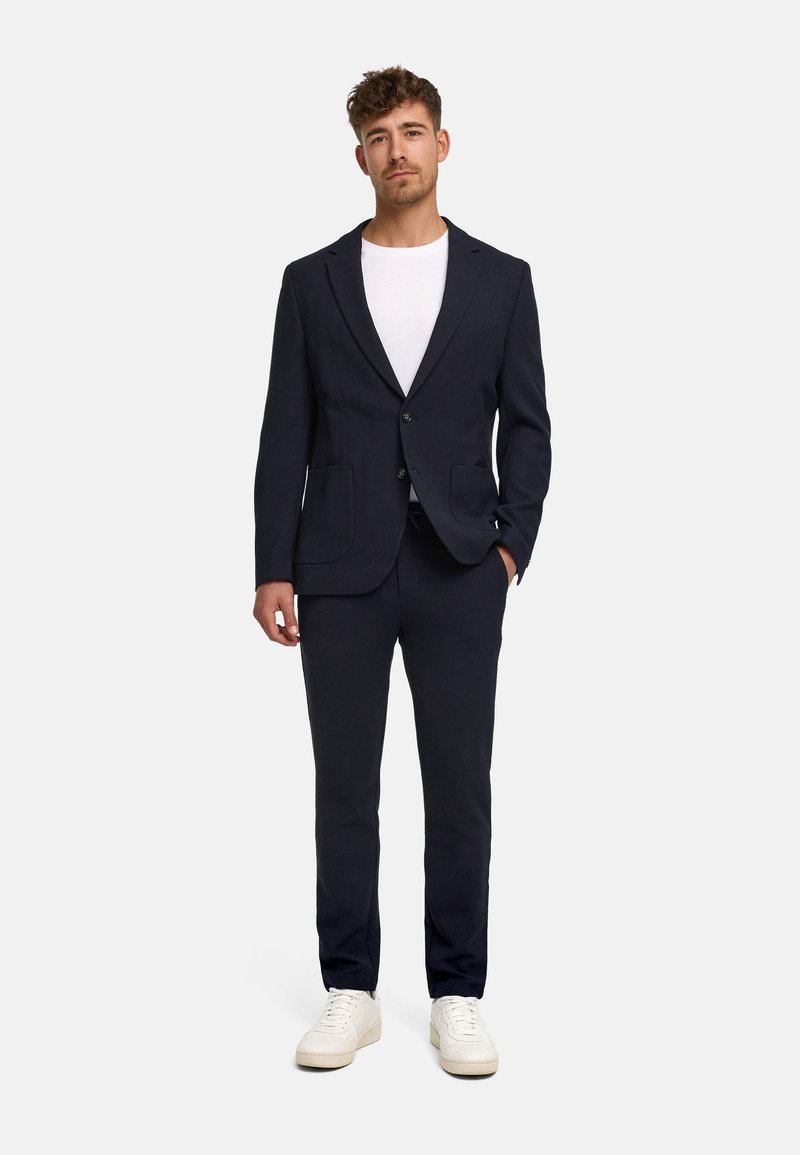 Homme debout, face à l’avant, habillé d’un costume noir avec une chemise blanche et des baskets blanches, une main dans la poche, fond neutre.