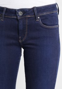 Jeans de mezclilla azul oscuro con un diseño ajustado. Presentan un estilo tradicional de cinco bolsillos, costuras en bronce y un cierre de botón metálico.