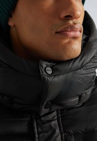 Schwarze Daunenjacke mit hohem Kragen und glänzender Oberfläche, ausgestattet mit einem sichtbaren Knopf und Logodetails am Hals.