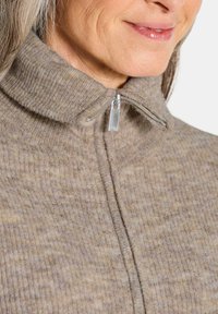Pull à côtes beige avec un col haut et une fermeture éclair en argent. Le tissu semble doux et texturé, présentant des variations de couleur subtiles.