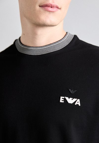 T-shirt noire avec un col rond rayé. Présente un logo "EVA" brodé en blanc et un petit emblème noir au-dessus. Texture lisse.