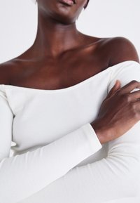 Off-the-shoulder vit ribbad topp med långa ärmar, med en texturerad tyg och en smal passform som framhäver halsringningen och axelområdet.