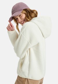 Crème kleurige fleece hoodie met een capuchon en zijzakken. Gecombineerd met een licht paarse corduroy pet. Zachte textuur en een losse pasvorm.