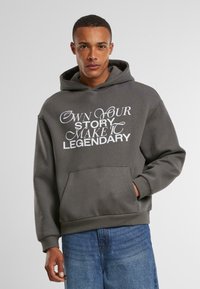Hombre con una sudadera con capucha gris oscuro y texto blanco "Haz tu propia historia, hazla legendaria" y jeans azules, de pie contra un fondo claro y liso.