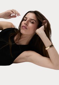 Top negro sin mangas, textura suave, usado por una persona con cabello largo y castaño. Pulsera de oro texturizada notable en la muñeca. Fondo neutral.
