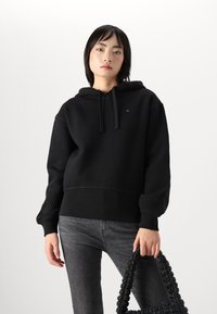 Hoodie in cotone nero con tasca frontale, cappuccio con cordino e dettaglio del logo. Il modello tiene una borsa nera texturizzata, indossando jeans grigi.