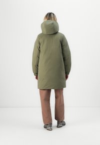 Olive green, knee-length puffer coat with a large hood, long sleeves, and elastic cuffs. Paired with brown pants and gray sneakers.