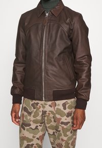 Man i en mörkbrun läderjacka med dragkedja och beige kamouflagedelar, som står mot en enkel ljus bakgrund.