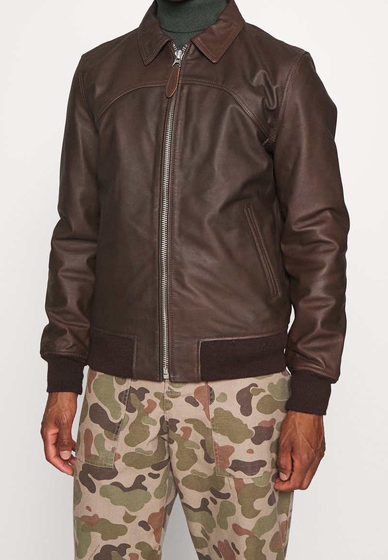 Uomo con giacca in pelle marrone scuro con zip e pantaloni beige a camouflage, in piedi contro uno sfondo chiaro e uniforme.