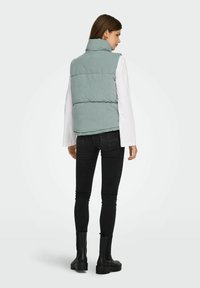 Donna che indossa un gilet imbottito senza maniche di colore azzurro chiaro, una camicia bianca a maniche lunghe, jeans skinny neri e stivali alla caviglia neri, in piedi di spalle.