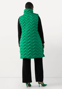 Mintgrön pufferväst med chevronmönster, hög krage och dragkedjestängning, ihopparad med svarta vida byxor och gröna klackar.