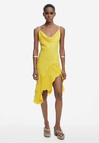 Robe jaune avec un col bénitier, un ourlet asymétrique et un détail de texture florale. Bretelles spaghetti et fente haute jusqu'à la cuisse. Finition en tissu lisse.