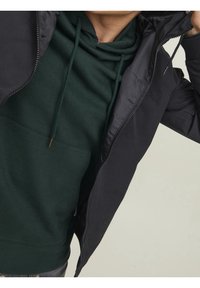 Svart zip-up jacka över en mörkgrön huvtröja. Jackan har en slät yta, medan huvtröjan har ett mjukt, borstat tyg och en framficka.