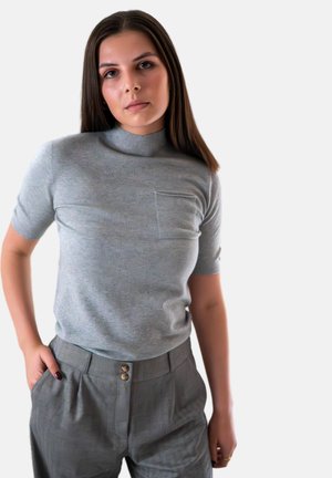 Vrouw met steil bruin haar die een grijze korte mouwen coltrui en hoge grijze broeken draagt, staande met één hand in haar zak.