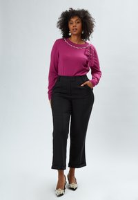 Pullover fucsia con perle decorative e dettaglio a fiocco abbinato a pantaloni neri a vita alta, sartoriali, e tacchi neri decorati.