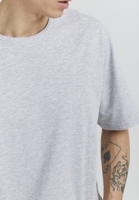 Gray cotton t-shirt with short sleeves, round neckline, and a soft texture. Includes an intricate tattoo on the arm featuring playing cards and figures.