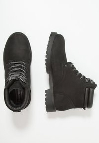 Černé semišové kotníkové boty s robustní podrážkou, zesílenými očky a černými a šedými tkaničkami. Obsahují polstrovaný límec pro pohodlí.