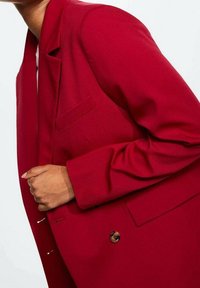 Personne portant un blazer cintré rouge vif avec un seul bouton visible, tenant le col près de la poitrine.