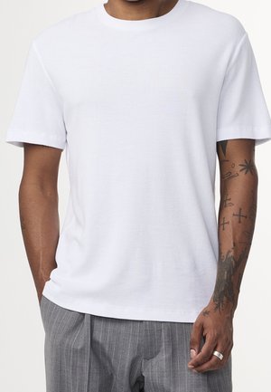 Hombre vistiendo una camiseta blanca lisa de manga corta y pantalones grises a rayas, con tatuajes visibles y un anillo de plata en la mano derecha.