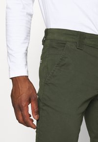 Olivgrön byxor med en slät textur, med sidofickor och en diskret logotyp. Matchat med en vit skjorta med långa ärmar.