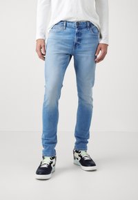 Hombre vestido con jeans ajustados azul claro, camisa blanca de manga larga y zapatillas negras y blancas con acentos verdes, de pie sobre un fondo liso.