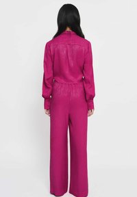 Fuksjowy kombinezon z wyraźnie zarysowanym kołnierzem, zapinanymi mankietami i szerokimi nogawkami. Gładka faktura i delikatne wzory na całej powierzchni.