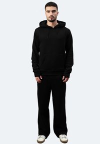 Schwarzer Kapuzenpullover und passende Hose aus weichem Material. Mit einer Fronttasche und Kordeln. Sneakers sorgen für einen lässigen Look.