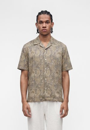 Νέος άνδρας με πλεγμένα μαλλιά που φοράει πουκάμισο με κοντά μανίκια με μπεζ σχέδια πασλέι και λευκό υφασμάτινο παντελόνι, στέκεται μπροστά σε λευκό φόντο.