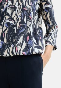 Bluza dugih rukava u tamnoplavoj boji s apstraktnim uzorkom koji uključuje ružičaste, bijele i plave elemente. Izrađena je od laganog, glatkog materijala.