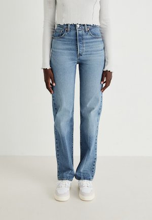 Personne portant un jean bleu clair à taille haute et jambes droites, un haut blanc à manches longues côtelé, et des baskets blanches, debout sur un fond blanc.