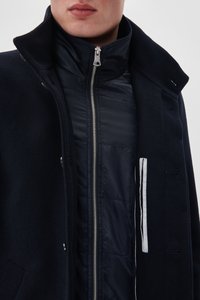 Mand iført en navy quilted jakke med lynlås lagd under en mørk uldfrakke med oprevet krave mod en ensfarvet baggrund.