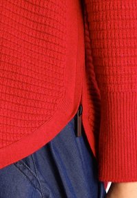 Rode gestructureerde cardigan met een geribde onderrand en ritsen aan de zijkant. De stof heeft een ruitpatroon, wat de tactiele aantrekkingskracht vergroot.