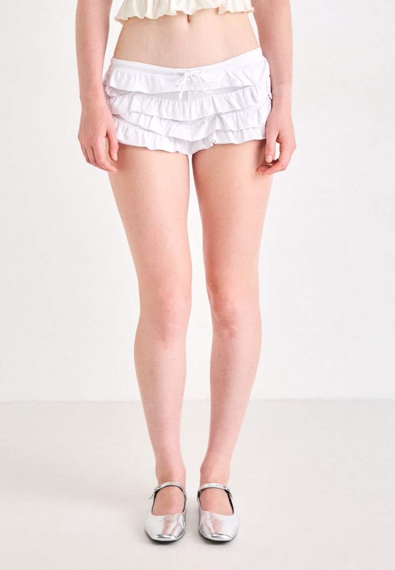 Shorts de bain blancs, à volants, avec une taille élastique et un détail de cordon de serrage. Associés à des chaussures plates en argent scintillant.
