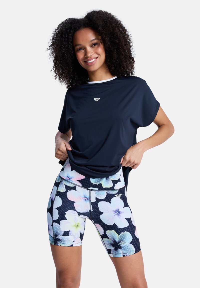 Kiharatukkainen nainen hymyilee, hänellä on päällään tumma lyhythihainen paita ja kukkakuosiset tiukat shortsit, ja hän säätää paitansa vyötäröltä.