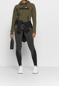 Olijfgroene hoodie met zwart logo, gecombineerd met donkergrijze en zwarte leggings met een kleurblokontwerp. Houdt een zwarte waterfles vast.