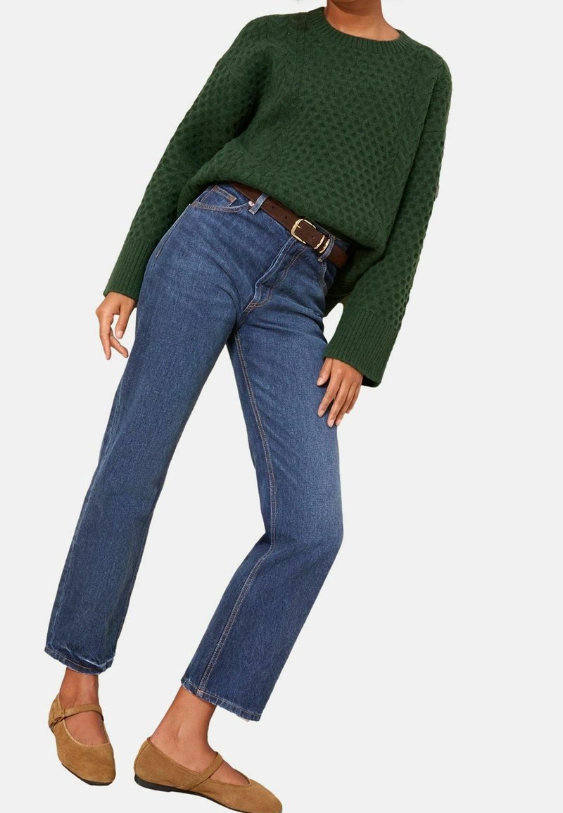 Personne portant un pull vert foncé texturé, un jean droit bleu avec une ceinture marron, et des chaussures plates en daim beige avec des sangles.